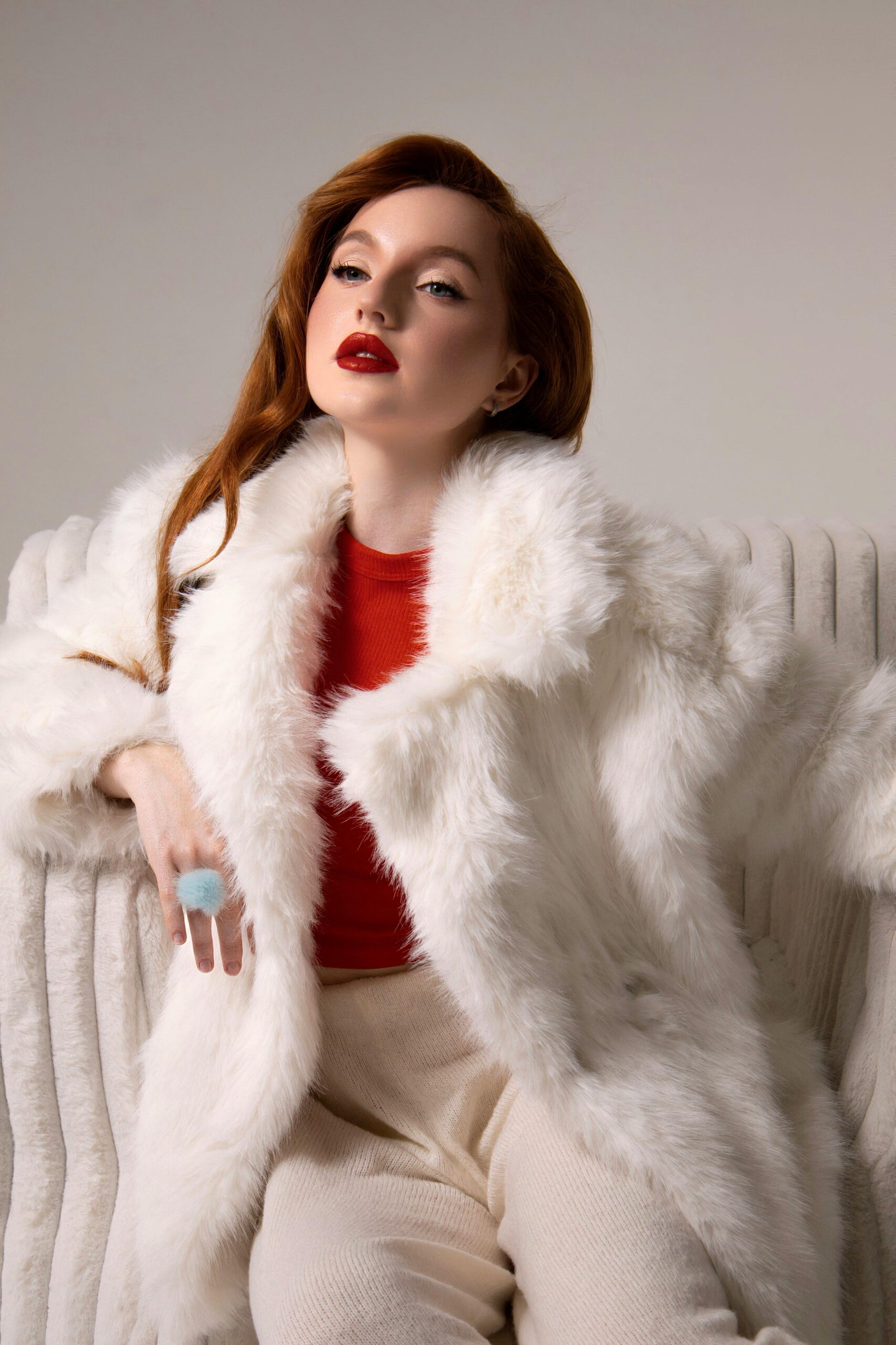 Home Stylish woman in luxurious white fur coat seated indoors with red lipstick and blouse.